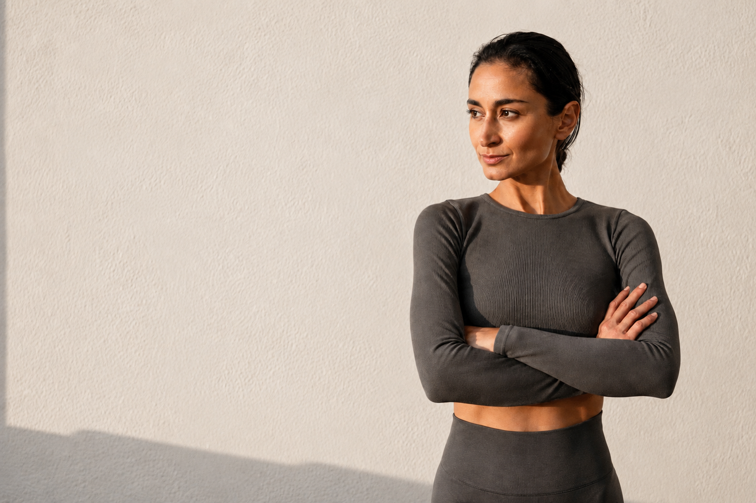 A woman in a dark neutral long-sleeve workout set standing with her arms crossed against a soft off-white wall.